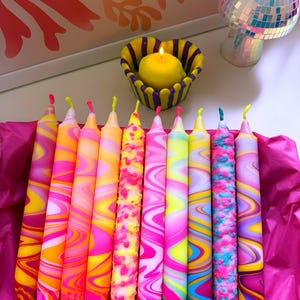 May include: A collection of ten taper candles featuring vibrant, multi-coloured marbled and textured wax patterns in shades of pink, orange, yellow, blue, and purple, arranged in a row on bright pink tissue paper with a lit yellow candle nearby.
