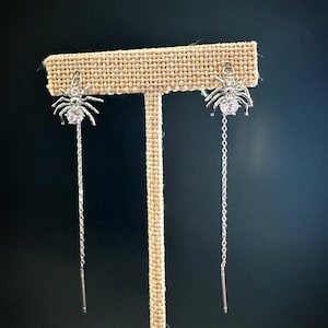 May include: A pair of silver-toned drop earrings featuring a spider design. Each spider has a clear, round faceted stone set in the abdomen and smaller stones on the head. A delicate silver chain hangs from the spider, ending in a thin metal post.