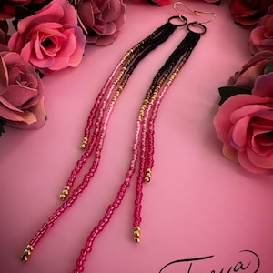 May include: Long, dangling earrings with black, gold, and pink beaded strands. The earrings have gold-colored hooks and are set against a pink background with roses. The word "Freya" is written in cursive.