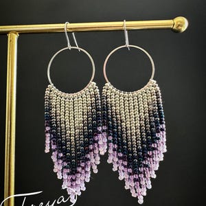 Pode incluir: Brincos pendentes com argolas de metal prateado e franjas de pequenas contas de vidro em degradê. As contas variam do prateado metálico no topo ao azul-marinho, roxo profundo e lavanda claro nas pontas inferiores.