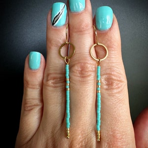 Puede incluir: Un par de pendientes largos de cuentas turquesa con aros dorados y cuentas doradas. Los pendientes cuelgan de la mano de una persona.