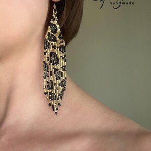May include: A pair of long, beaded dangle earrings with a black and gold leopard print pattern. The earrings are handmade and feature a fringe design.