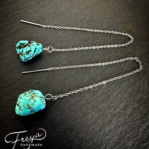 May include: A pair of turquoise stone earrings with silver chains. The stones are a light blue color with brown veins. The chains are delicate and have a simple design.