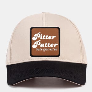 May include: A beige baseball cap with a black brim. The cap has a brown square patch with white text that reads "Pitter Patter let's get at 'er".