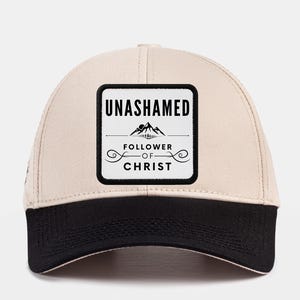 Peut inclure: Casquette de baseball beige avec une visière noire. Le devant de la casquette présente un patch carré noir avec du texte blanc qui dit "UNASHAMED FOLLOWER OF CHRIST" avec un graphique de montagne.