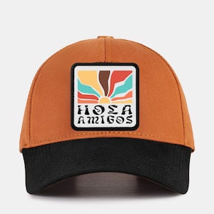 May include: A brown and black baseball cap with a colourful patch that says "Hola Amigos" in a retro font.