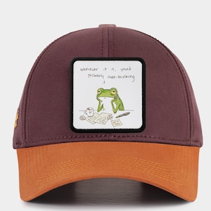 May include: A maroon and orange baseball cap with a square patch featuring a cartoon frog and the text "whatever it is, you're probably over-thinking it."