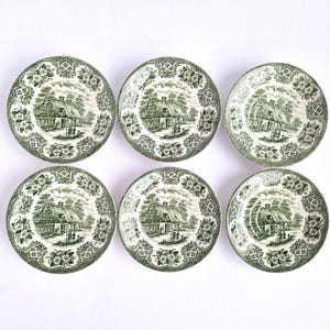 Può includere: Un set di sei piatti rotondi in ceramica con un motivo a trasferimento verde e bianco. Ogni piatto presenta un'illustrazione centrale di un cottage dal tetto di paglia con figure, circondato da un bordo decorativo di fiori e forme geometriche.