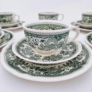 Może przedstawiać: Zestaw białych filiżanek do herbaty, spodków i talerzy w stylu vintage z zielonym kwiatowym wzorem. Filiżanki mają zaokrąglony kształt z uchwytem. Talerze i spodki mają falisty brzeg. Kwiatowy wzór zawiera kwiaty i liście.