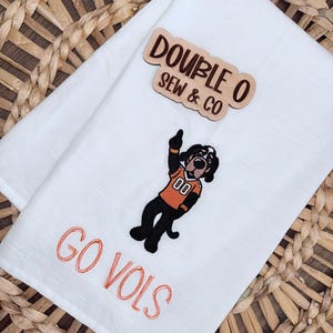 May include: A white kitchen towel featuring an embroidered graphic of a dog wearing an orange jersey with the number 00, pointing upward. Below the dog, the text GO VOLS is embroidered in orange. A wooden sign reads DOUBLE O SEW & CO.