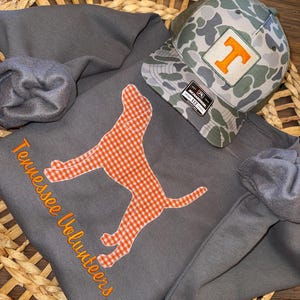 May include: A grey sweatshirt with orange embroidery reading "Tennessee Volunteers" and a gingham dog silhouette. A camouflage baseball cap with an orange "T" patch rests on top. The items are displayed on a woven basket.