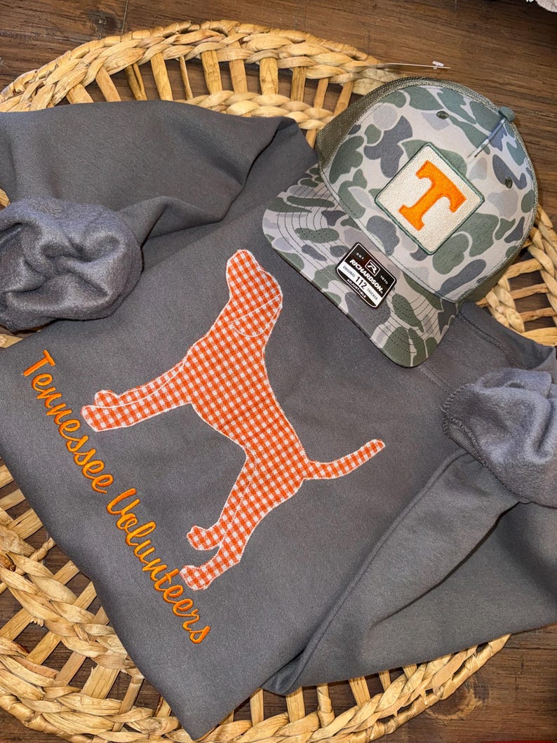 Op de afbeelding: Een grijze sweatshirt met oranje borduursel met de tekst "Tennessee Volunteers" en een geruite hondenomtrek. Een camouflage baseballcap met een oranje "T" patch ligt erbovenop. De items worden gepresenteerd in een gevlochten mand.
