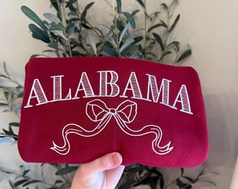 May include: A maroon sweatshirt with the word "ALABAMA" embroidered in white script, along with a bow and ribbon design. The sweatshirt is held up in front of a background of green leaves.