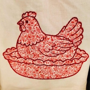 May include: Embroidered hen design on cream fabric. The hen features a red floral pattern and red thread outline. The hen sits on a red, scalloped base, creating a charming, rustic aesthetic. The design is reminiscent of country crafts.