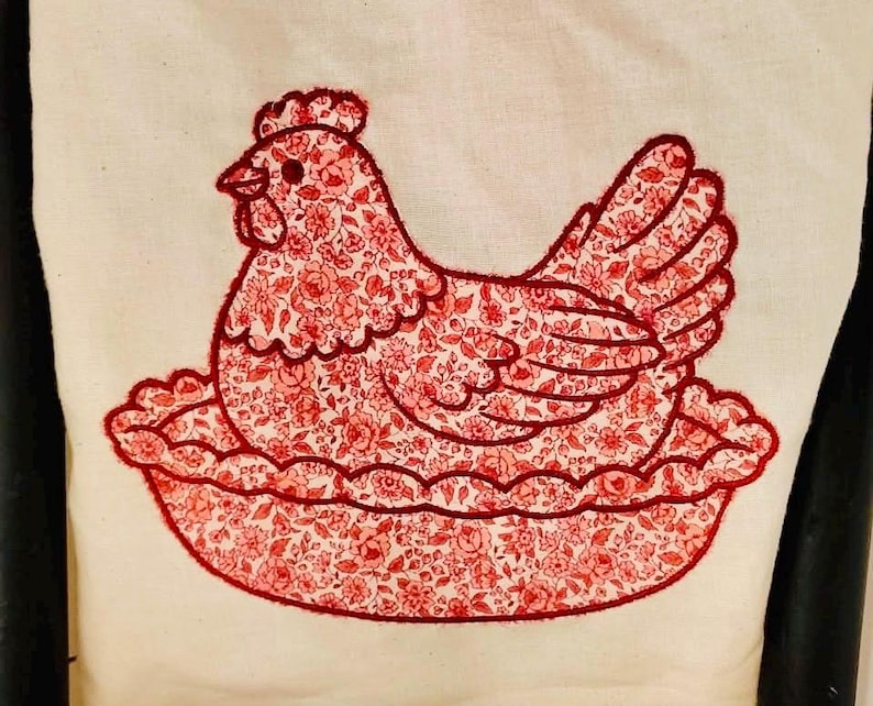 May include: Embroidered hen design on a cream-colored fabric. The hen is filled with a red floral pattern and outlined in red thread. The hen is sitting on a red scalloped base, creating a charming, rustic aesthetic.