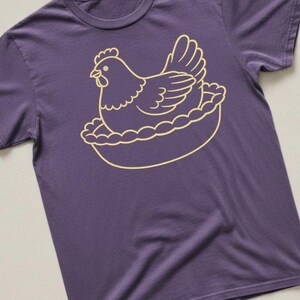May include: A purple t-shirt featuring a gold-colored illustration of a hen sitting in a nest. The hen is depicted in a simple, cartoon-like style, with detailed feathers and a comb. The nest has a scalloped edge.