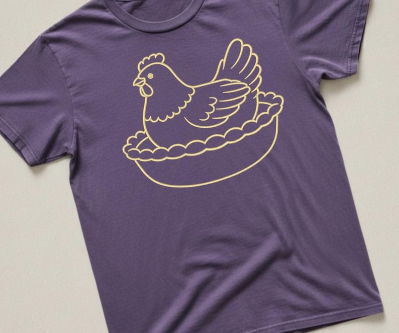 May include: A purple t-shirt featuring a gold-colored illustration of a hen sitting in a nest. The hen is depicted in a simple, cartoon-like style, with detailed feathers and a comb. The nest has a scalloped edge.