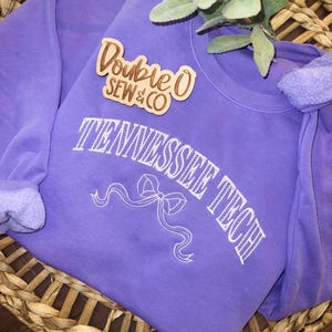May include: A lavender sweatshirt with the text "TENNESSEE TECH" in white script and a bow graphic. A wooden pin with the text "Double O SEW & CO" is pinned on the sweatshirt. The sweatshirt is displayed in a woven basket.