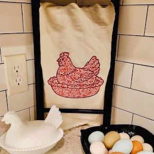 May include: A cream-colored kitchen towel with a red floral hen design hangs on a black ladder. A white ceramic hen-shaped egg holder and a black bowl of eggs are on the counter. The eggs are various colors, including blue, brown, and white.