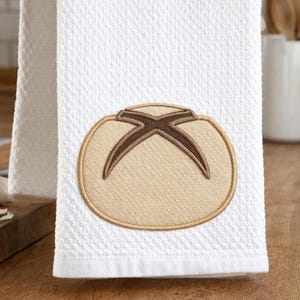 Puede incluir: Toalla de cocina blanca con tejido de gofre. Presenta un diseño bordado de una hogaza de pan en beige y marrón. La toalla está doblada y colgada, con una tabla de cortar de madera y ajo en el fondo.