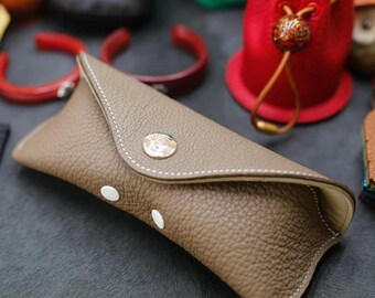 May include: A brown leather glasses case with a white stitching detail and a silver button closure. The case is folded shut and has a soft, textured surface.