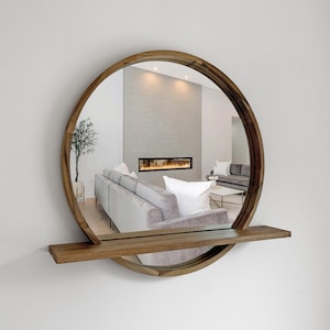 May include: A round wooden mirror with a built-in shelf. The mirror's frame is made of light brown wood. The mirror reflects a living room with a gray sofa, white pillows, and a fireplace. The shelf is rectangular and made of the same wood as the frame.