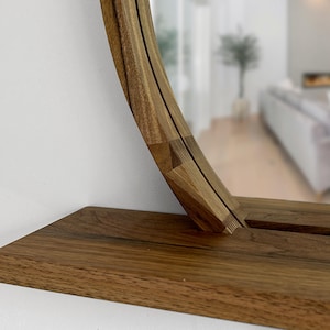May include: A wooden, round mirror with a shelf. The mirror frame and shelf are made of a light brown wood. The mirror is reflecting a room with a white wall and a couch.