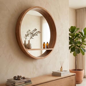 Puede incluir: Un espejo de pared redondo de madera con un estante horizontal integrado. El marco del espejo es de madera clara y contiene dos botellas pequeñas de vidrio ámbar y un jarrón pequeño con ramas verdes reflejadas en la superficie del estante.