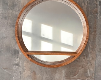 May include: A round wall mirror featuring a thick, natural-toned wooden frame and a horizontal wooden shelf integrated across the lower third of the glass surface. The mirror reflects light, showing a bright, rectangular window pattern.