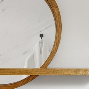 May include: A round wooden framed mirror with a shelf. A white towel hangs from a black hook on the wall. The mirror and shelf have a natural wood grain finish. The wall is white with a marble-like texture.