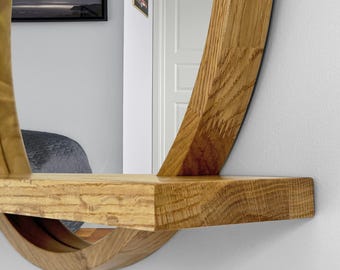 May include: A circular wall mirror featuring a light brown oak wood frame with a prominent natural grain pattern. A horizontal wooden shelf is integrated into the lower section of the circular frame, extending outward from the mirror surface.