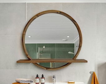 May include: A round wooden-framed mirror with an integrated horizontal shelf across the bottom third. The mirror reflects a bathroom interior with green walls and a white ceiling. The frame features a natural wood grain finish.