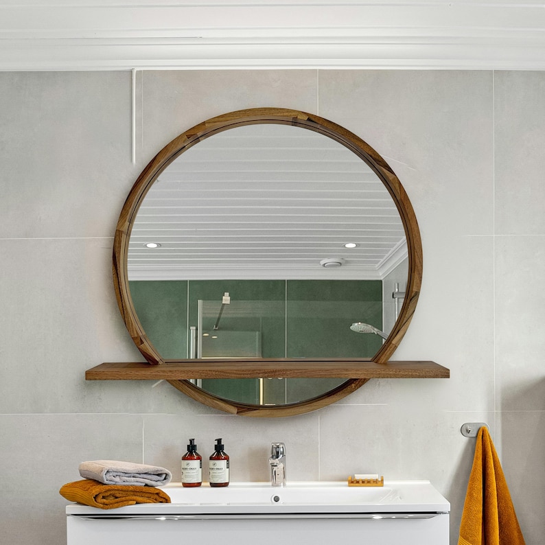 May include: A round wooden framed mirror with a built-in shelf. The mirror is mounted on a light gray tiled wall above a white sink. The bathroom features a modern design with a minimalist aesthetic.