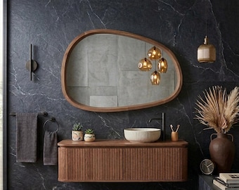 May include: A wall-mounted wooden vanity with a slatted front, topped with a ribbed ceramic vessel sink and a matte black faucet. Above it hangs an organic-shaped wooden mirror reflecting a cluster of amber glass pendant lights.