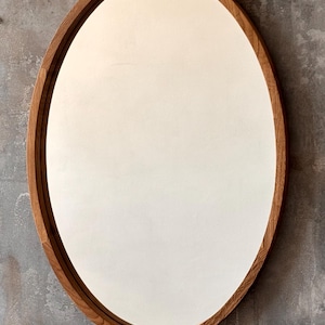 May include: Oval mirror with a light brown wooden frame. The mirror's surface is white and reflects light. The mirror is mounted on a grey textured wall.