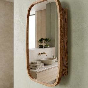 May include: A wall-mounted medicine cabinet featuring a rectangular mirror with rounded corners and a thick, natural wood frame with a visible grain pattern. The cabinet is mounted on a light green wall, reflecting a bathroom vanity with a sink.