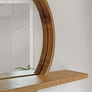 May include: A round wooden mirror with a shelf. The mirror has a natural wood grain finish and a curved frame. The shelf is made of the same wood and is rectangular. The mirror is mounted on a white wall.