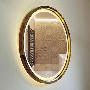 May include: Oval-shaped illuminated mirror with a wooden frame. The frame has a mosaic-like pattern of various wood tones. The mirror has a warm, soft glow around the edge, creating a modern and stylish look. The mirror is mounted on a white wall.