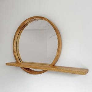Pode incluir: Um espelho redondo de madeira com uma prateleira. O espelho tem uma moldura circular de madeira castanha clara. A prateleira retangular também é de madeira. O espelho está montado numa parede branca. O diâmetro do espelho é de cerca de 60 cm.