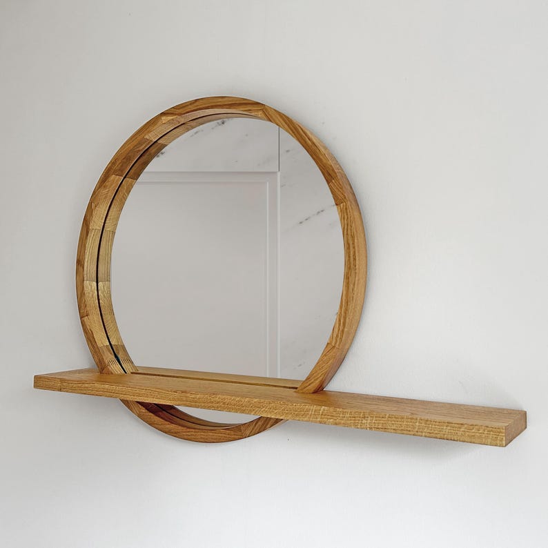 May include: A round wooden mirror with a shelf. The mirror has a circular frame made of light brown wood. The shelf is rectangular and made of the same wood. The mirror is mounted on a white wall.