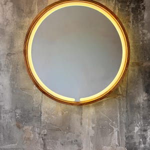 May include: A circular mirror featuring a wooden frame and a warm, yellow LED light. The mirror is mounted on a textured grey wall, with a wooden surface below. The design of the mirror is modern and minimalist, suitable for various interior styles.