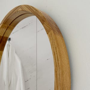 May include: A close-up of a wooden-framed, oval mirror. The frame is made of light-colored wood with a natural grain. The mirror reflects a white bathrobe hanging on a hook. The background is a white wall.