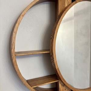 May include: A circular wooden wall unit featuring a natural light brown wood grain finish. The structure includes a round mirror on the right side and two horizontal shelves on the left side, all contained within a large circular wooden frame.
