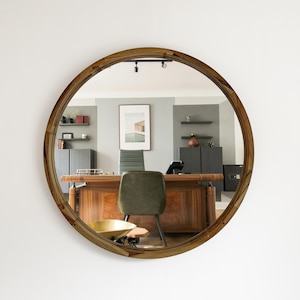 May include: A round wooden mirror with a decorative frame. The mirror reflects an office setting with a desk, chair, and artwork. The frame has a natural wood grain pattern, adding a touch of warmth to the space.