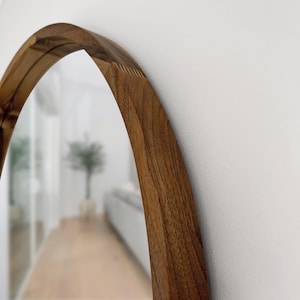 May include: A wooden-framed, arched mirror. The frame is made of a light brown wood with visible grain. The mirror reflects a blurred interior scene with a hallway and a plant.