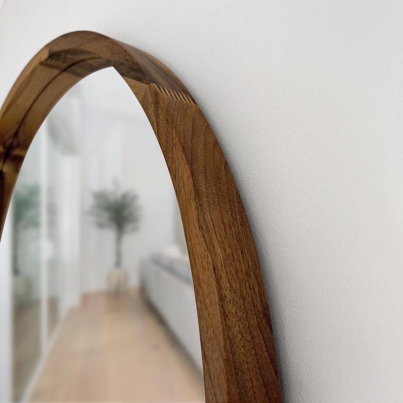 May include: A wooden-framed, arched mirror. The frame is made of a light brown wood with visible grain. The mirror reflects a blurred interior scene with a hallway and a plant.