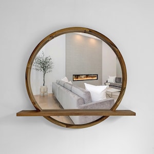 May include: A round wooden mirror with a built-in shelf. The mirror has a natural wood grain finish and reflects a living room scene with a fireplace, sofa, and a small potted tree. The shelf is rectangular and made of the same wood.