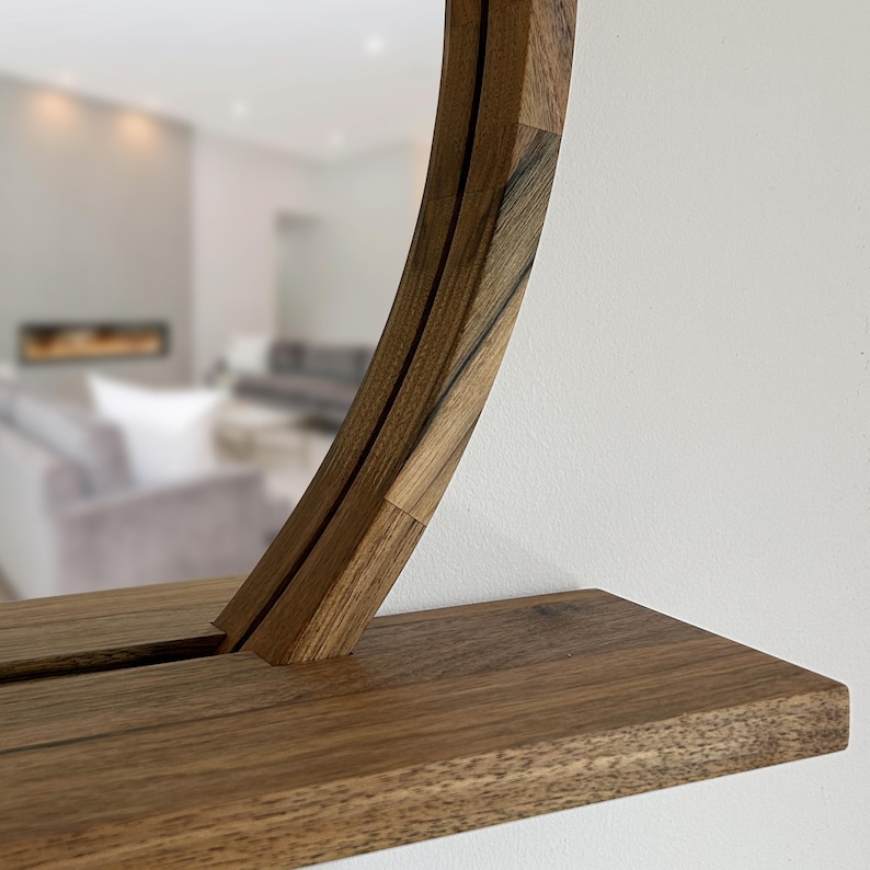 May include: A round wooden mirror with a shelf. The mirror's frame and shelf are made of dark brown wood. The frame has a unique, curved design. The mirror is mounted on a white wall, with a blurred living room in the background.
