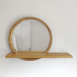 May include: A round wooden mirror with a shelf. The mirror has a natural wood finish and a circular frame. The shelf is rectangular and extends from the bottom of the mirror. The design is minimalist and modern, suitable for a bathroom or entryway.