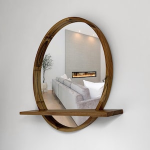 May include: A round wooden mirror with a built-in shelf. The mirror has a natural wood finish and a curved design. The reflection shows a living room with a sofa, fireplace, and a small tree in a pot. The mirror is mounted on a white wall.